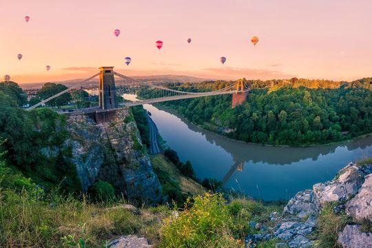 Many Hot Air Balloons drift towards Clifton Suspension Bridge in Bristol at sunrise. The bridge spans the River Avon gorge.